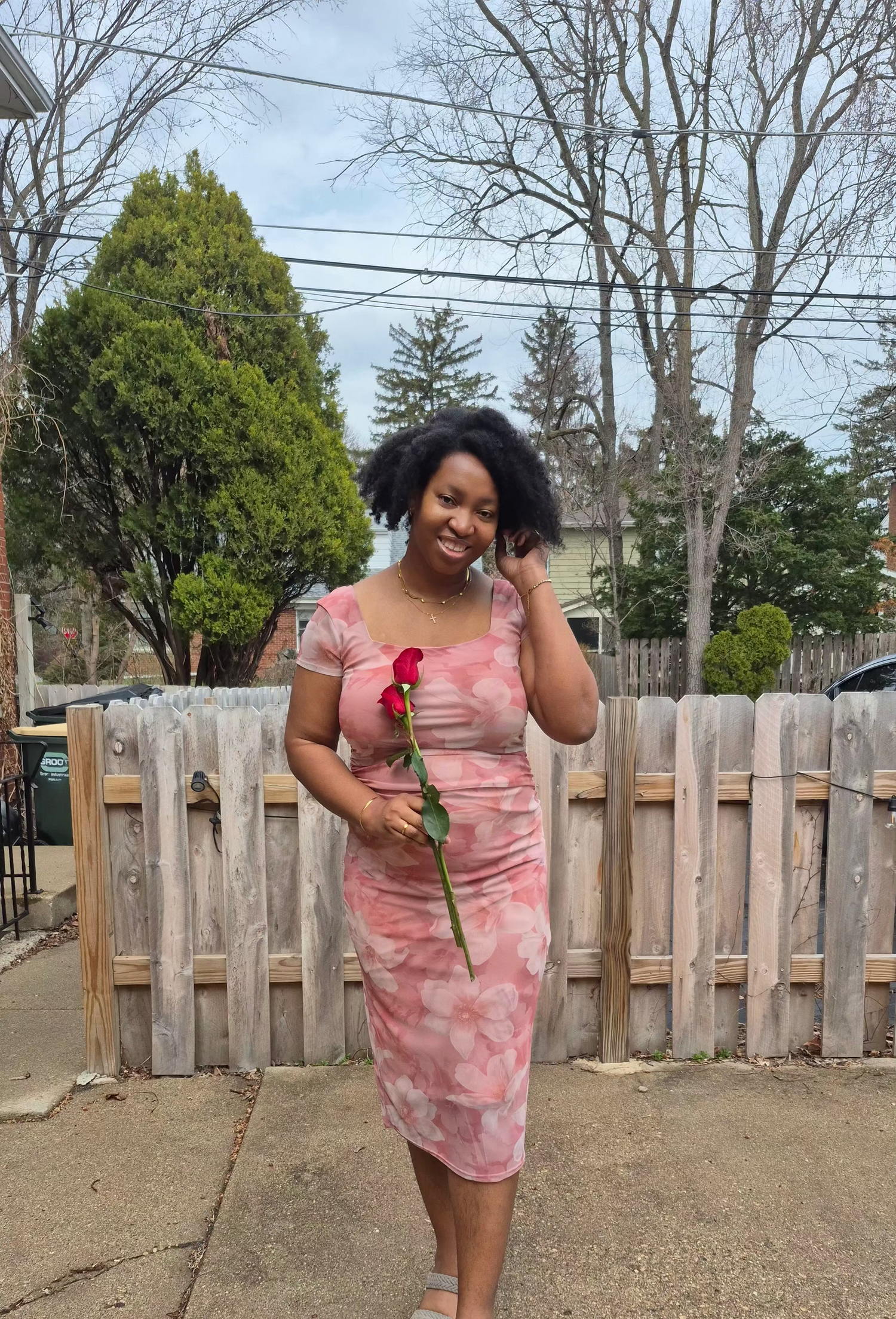 Woman in a pink dress holding a red rose on a sidewalk with trees and a wooden fence in the background.